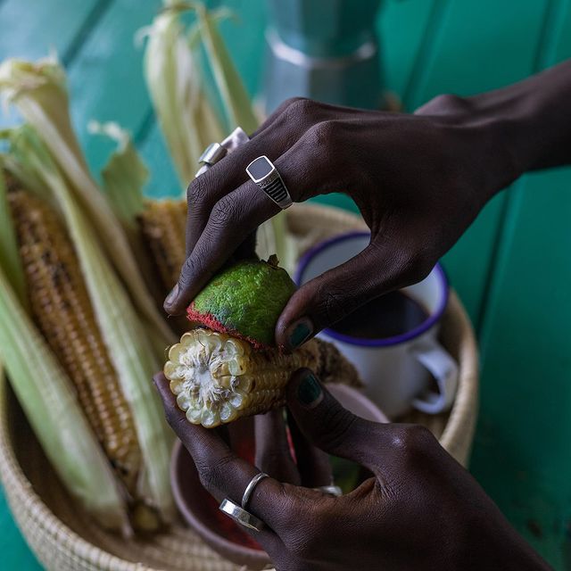 A picture of a hand holding roasted maize coated with pepper and lemon, with other roasted maize in a basket